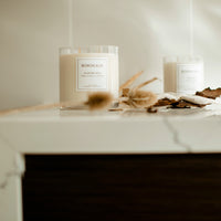 Thumbnail for Small Deluxe Harvest | Leather & Sandalwood | 3 Wicks | Glass Vessel sitting on a kitchen bench | Bordeaux Candles