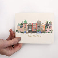 Hand holding a card with a colorful illustration of buildings and 'Happy New Home' text on a white background