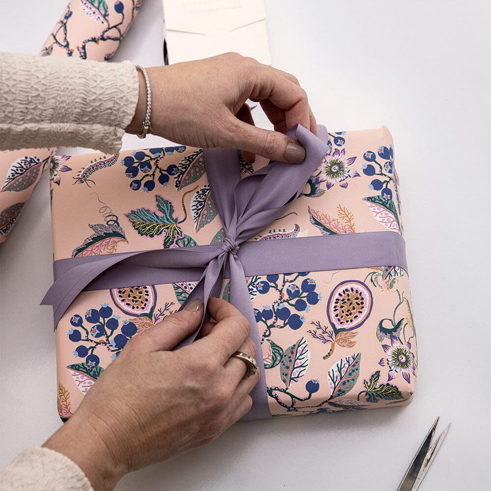 Person wrapping a gift with floral paper and a purple ribbon on a white surface.