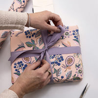 Person wrapping a gift with floral paper and a purple ribbon on a white surface.